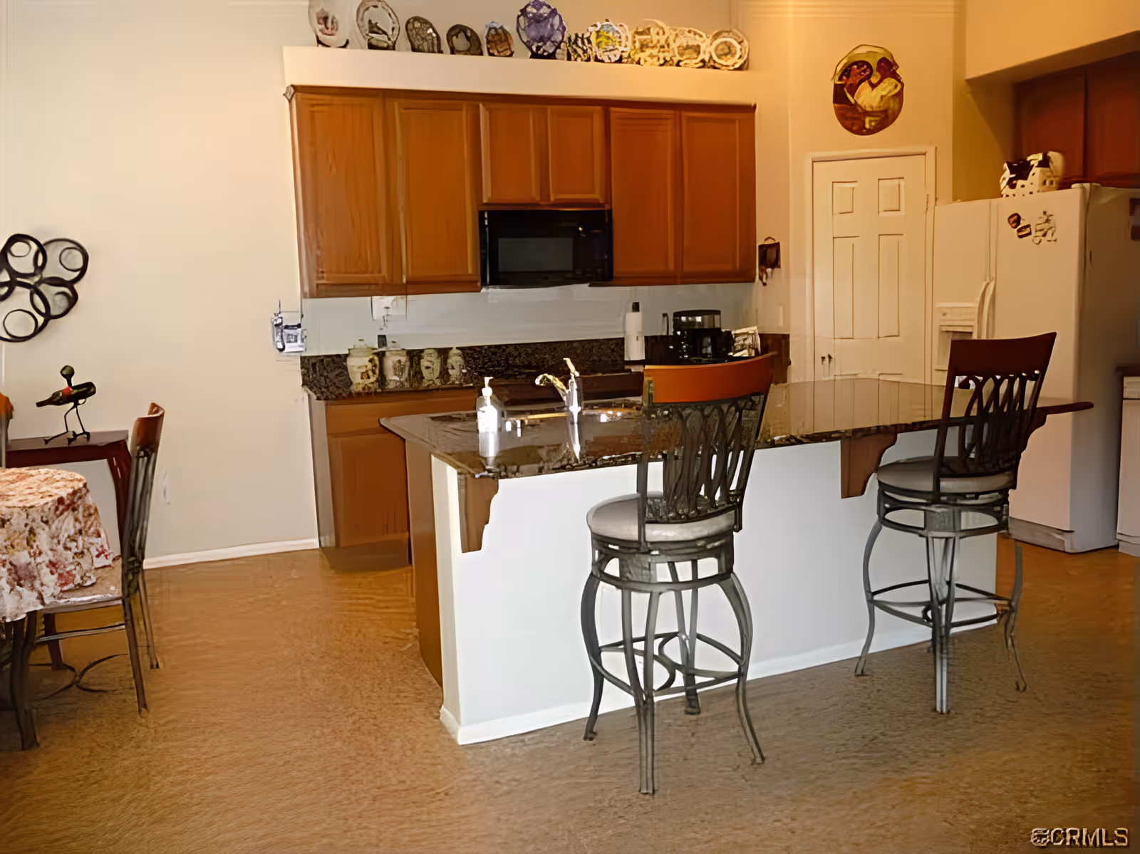 Open kitchen with a granite island and two bar stools, wooden cabinets, a refrigerator, and an adjacent dining table.