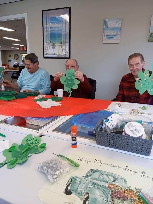Three elderly men sitting at a table covered with a red tablecloth, engaging in a craft activity making green shamrock decorations. The table has various craft supplies including glue sticks, googly eyes, and paper. The room has light-colored walls with framed pictures, one showing a beach scene and another with a boat.