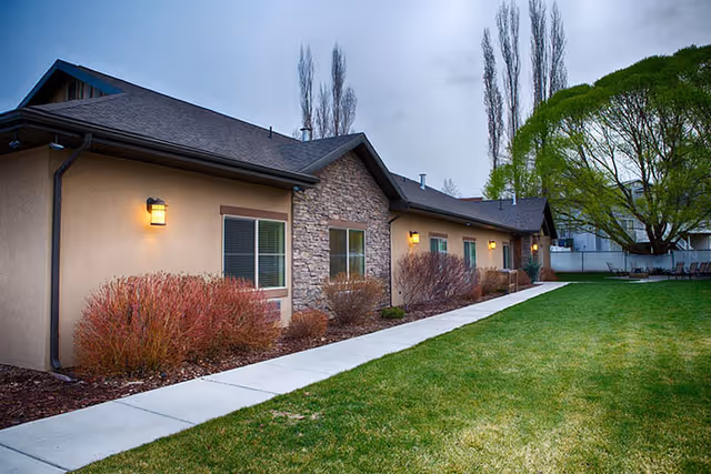 Exterior view of a single-story assisted living facility building with beige walls and stone accents. The building is bordered by a concrete walkway and landscaped with bushes and a large tree on a grassy lawn under a cloudy sky.
