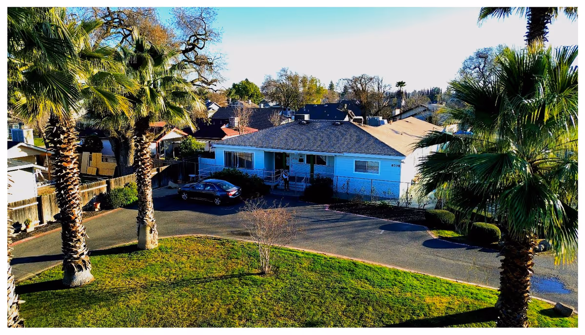Exterior view of a single-story residential building with a dark shingled roof, surrounded by palm trees and greenery. A car is parked in the driveway in front of the building, and a person is standing near the entrance. The sky is clear and blue.