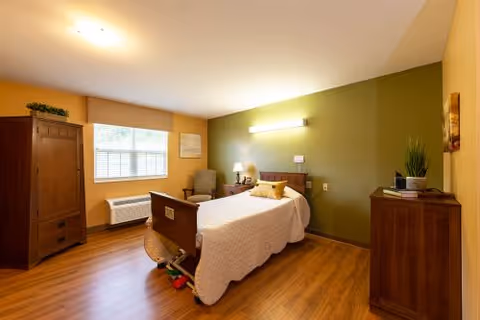 A well-lit bedroom in a skilled nursing facility featuring a single hospital-style bed with white bedding and a yellow pillow. The room has wooden flooring, a green accent wall behind the bed, a wooden wardrobe, a wooden dresser with a plant and books, a comfortable armchair, and a window with blinds letting in natural light.