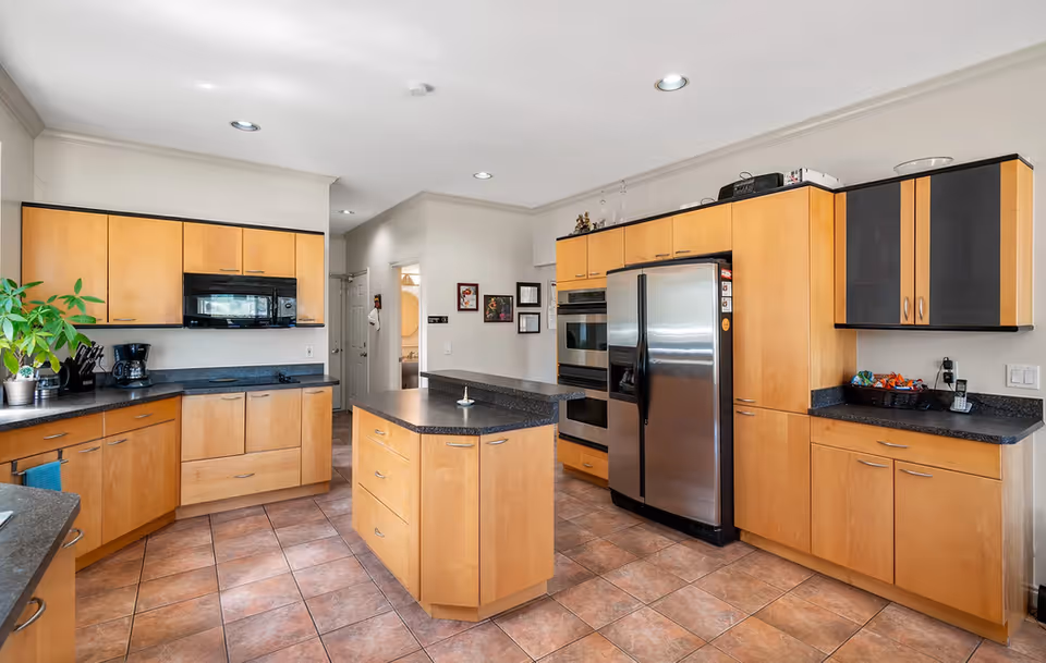 A modern kitchen with light wood cabinets, black countertops, and tiled floor. The kitchen features a central island, a stainless steel refrigerator, built-in oven, microwave, coffee maker, and a few decorative items including a plant and framed pictures on the wall.