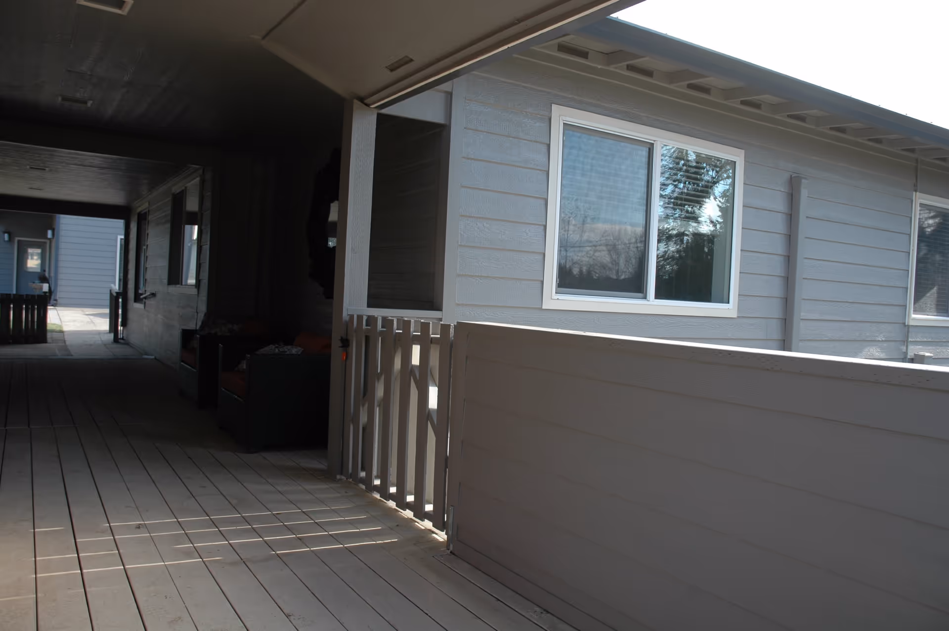 Covered outdoor walkway with wooden flooring and gray siding walls, featuring a small gate and windows reflecting trees outside. There is some seating visible in the shaded area along the wall.