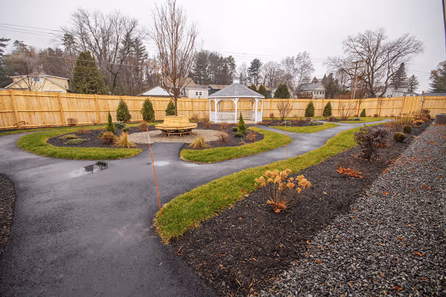 Fenced outdoor courtyard with winding paved paths, landscaped beds, a central round bench and a white gazebo.