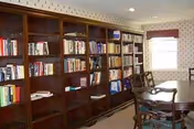 A cozy interior room featuring a large wooden bookshelf filled with books along one wall, a round wooden table with chairs, and a window with a red valance allowing natural light to enter.