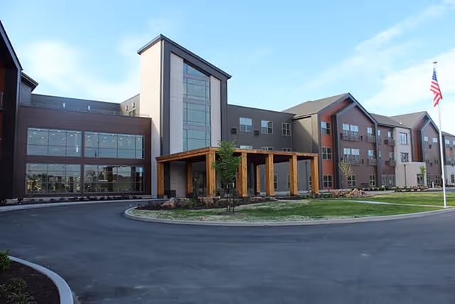 Exterior view of a modern three-story senior living facility building with large windows, a covered entrance with wooden beams, landscaped grounds, and an American flag on a flagpole.
