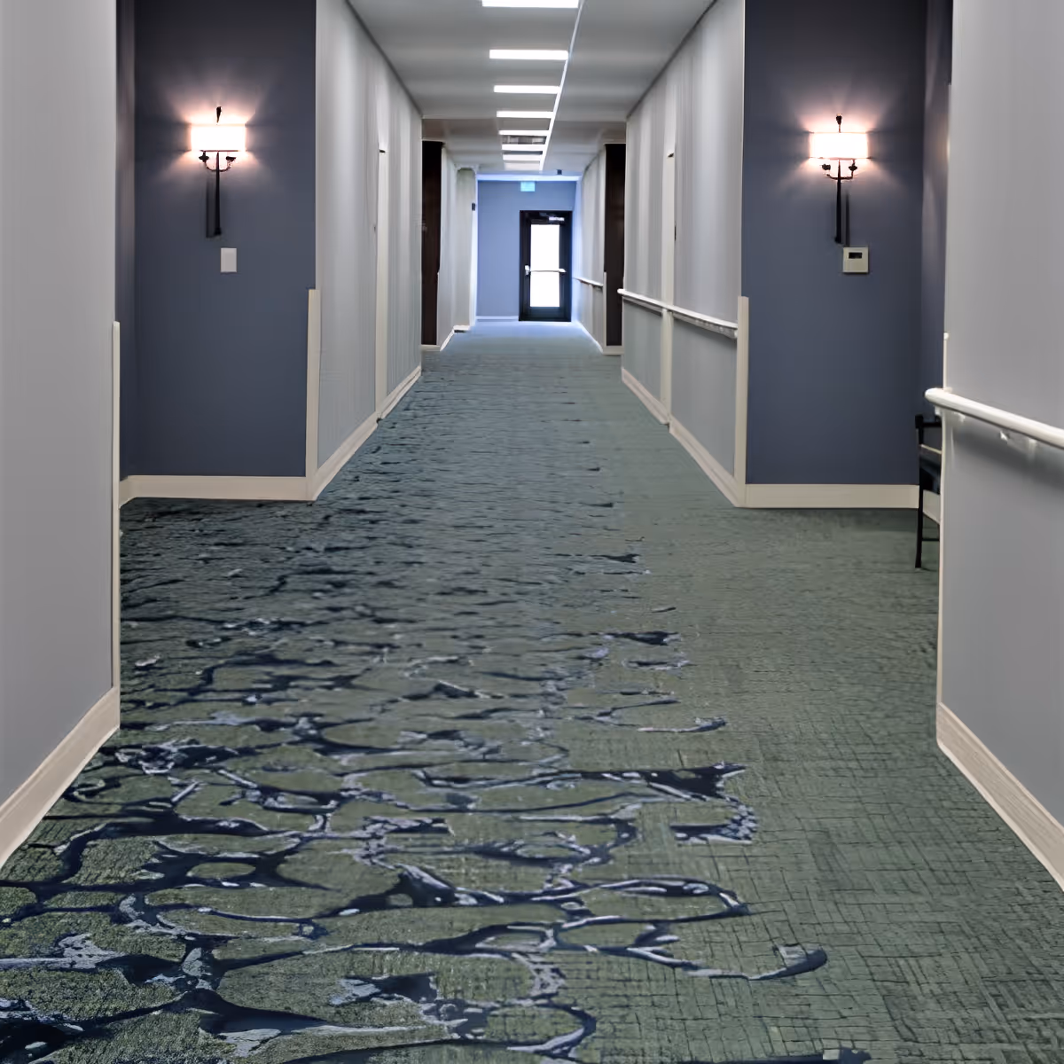 A long indoor hallway with blue and gray walls, blue patterned carpet, wall-mounted light fixtures, and handrails on both sides. The hallway has several doors along the walls and a glass exit door at the far end.