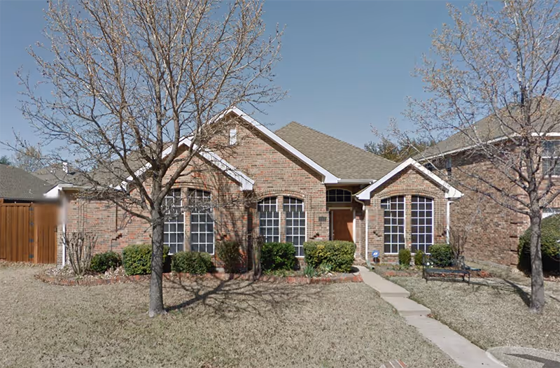 Front exterior view of a single-story brick house with a gabled roof, multiple large windows, a wooden front door, and a concrete pathway leading to the entrance. The yard has two leafless trees and some bushes near the house under a clear blue sky.