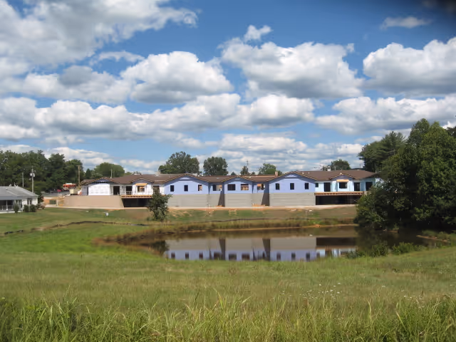 A row of buildings under construction with blue sheathing on the exterior walls, situated behind a small pond and grassy field under a partly cloudy sky.