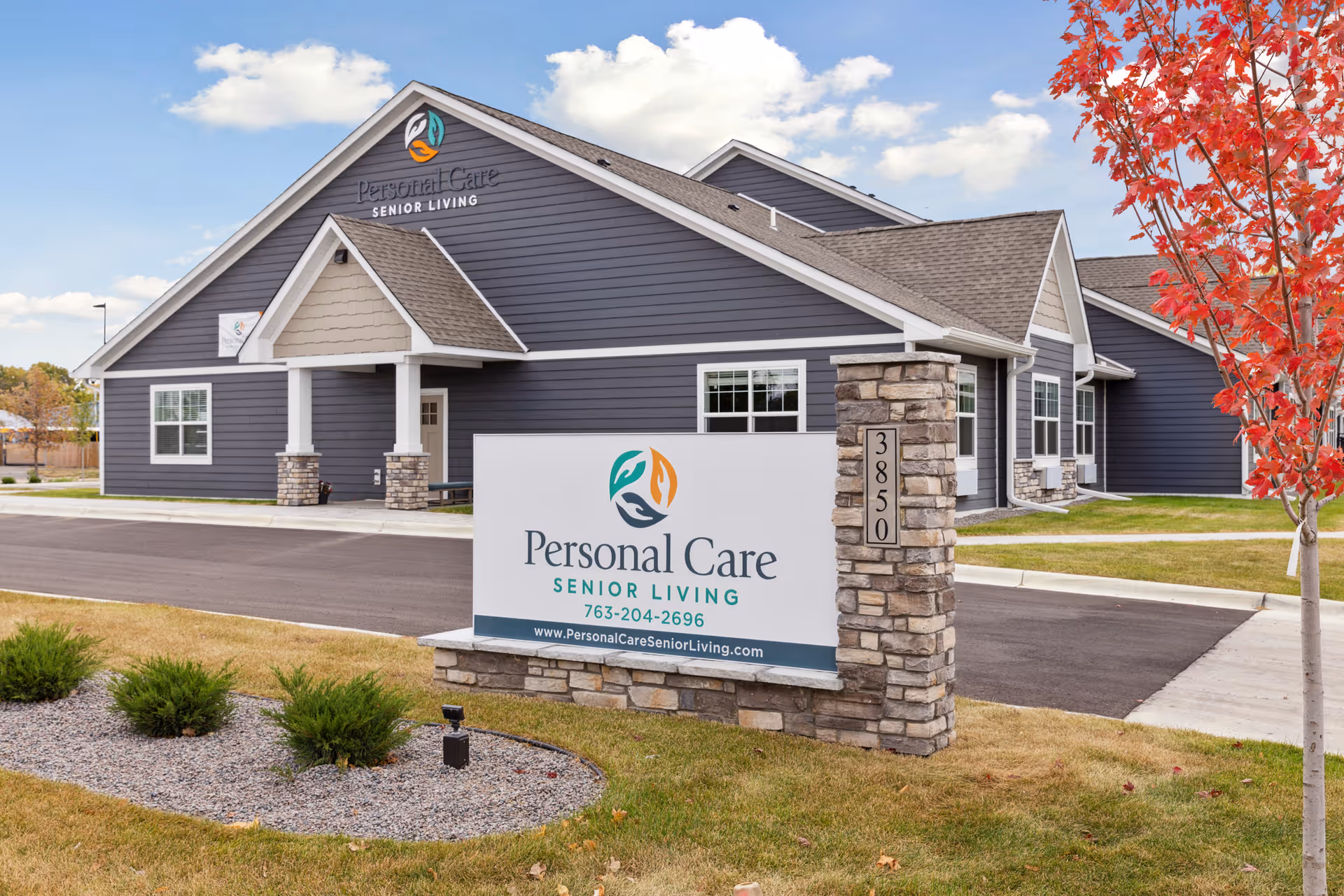 Exterior view of Personal Care Senior Living facility in Columbia Heights, showing a modern building with gray siding and stone accents. A large sign in front displays the facility's name, phone number, and website. There is a small landscaped area with bushes and a tree with red leaves beside the sign.