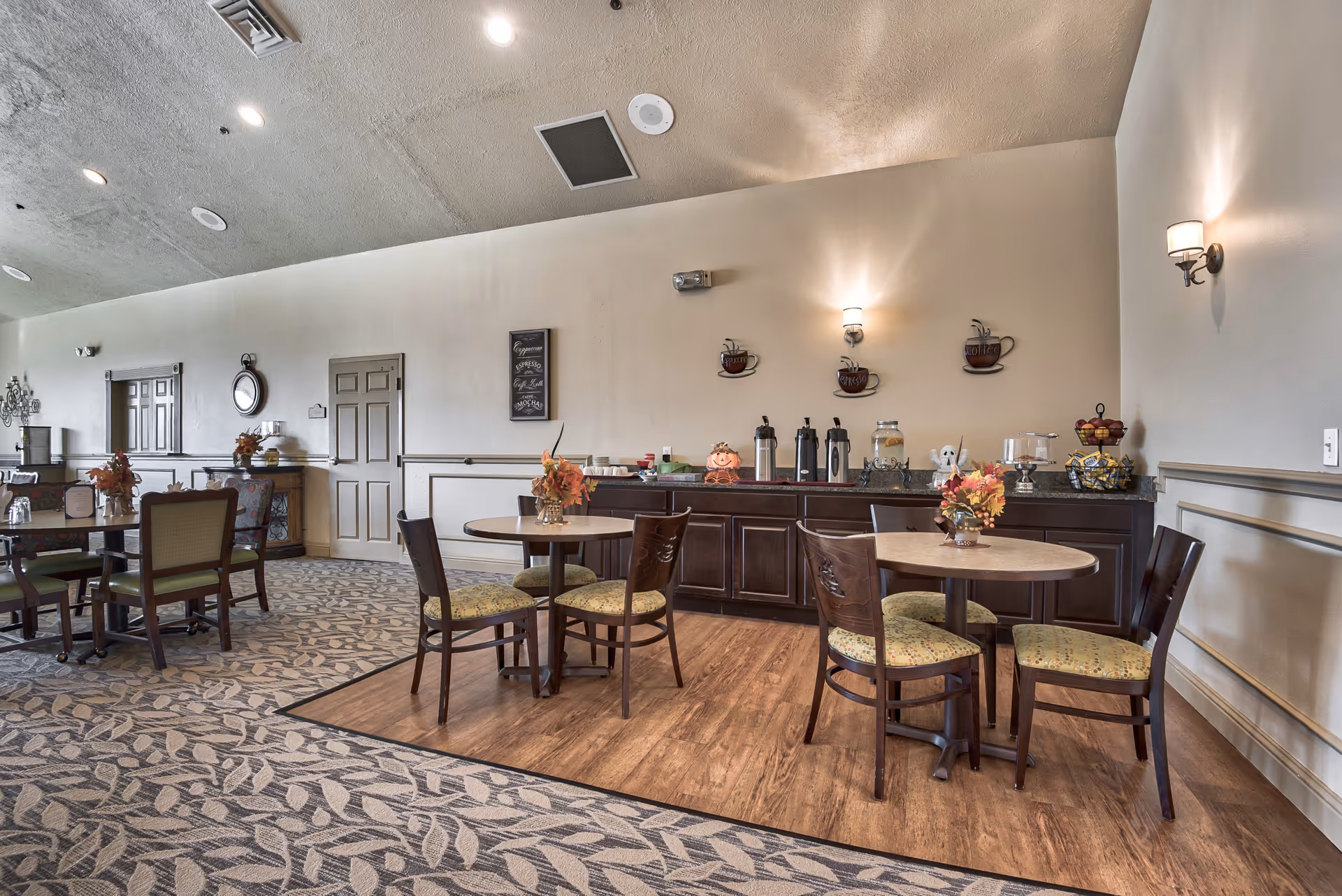 A cozy dining area in Oak Pointe Assisted Living featuring round tables with chairs, floral centerpieces, and a coffee station against the wall with coffee-themed decorations and warm lighting.