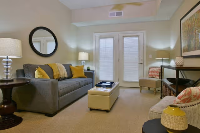 A cozy living room with a gray sofa adorned with yellow and gray pillows and a yellow throw blanket. There is a round mirror on the wall above the sofa, two table lamps on either side, a beige ottoman in the center with a tray on top, a beige armchair with a patterned pillow, and a console table with decorative items and baskets underneath. French doors with blinds are at the back of the room.
