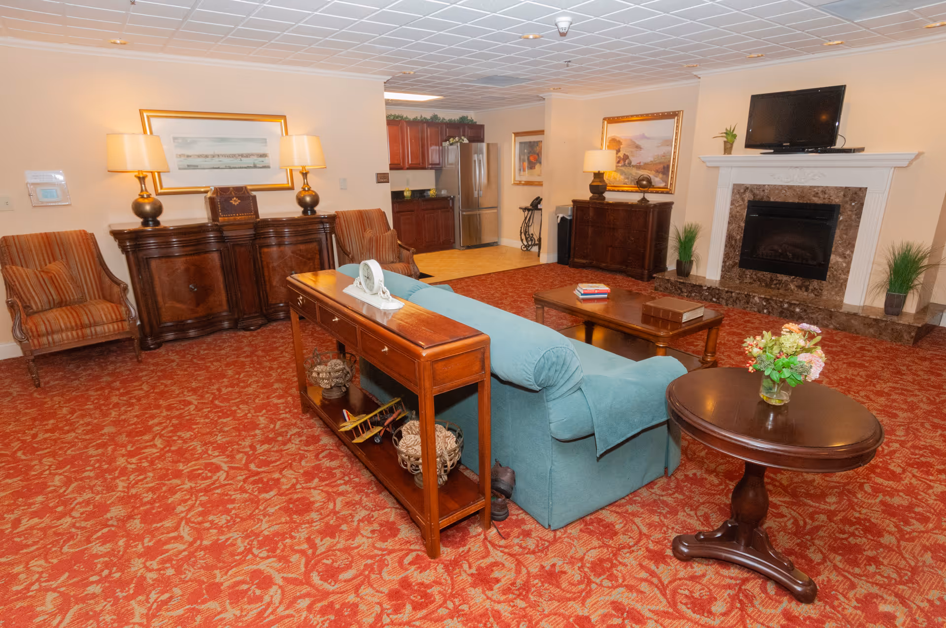 A cozy living room area with a red patterned carpet, a teal sofa, two striped armchairs, wooden side tables, and a coffee table with books. The room features a fireplace with a TV mounted above it, two framed paintings on the walls, and a small kitchenette area with a refrigerator and cabinets in the background.