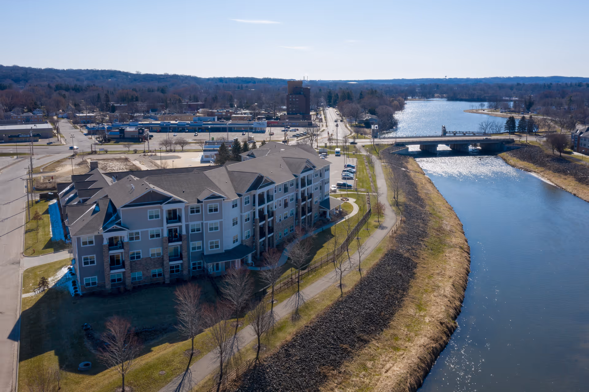 Aerial view of a multi-story residential building next to a river with a nearby bridge, parking lot, and tree-lined walking path.