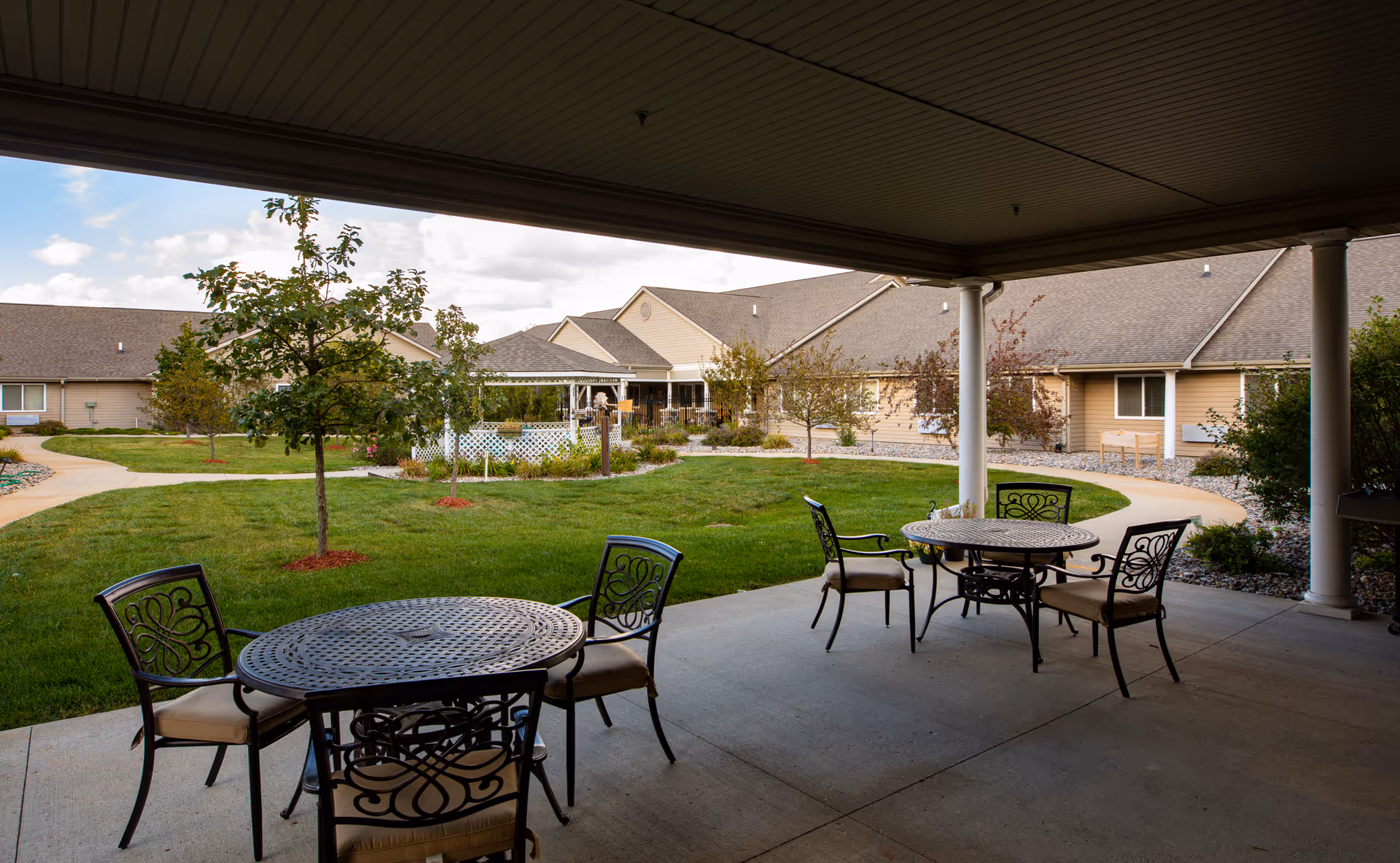 Covered patio with metal tables and chairs overlooking a landscaped courtyard and single-story assisted living buildings.