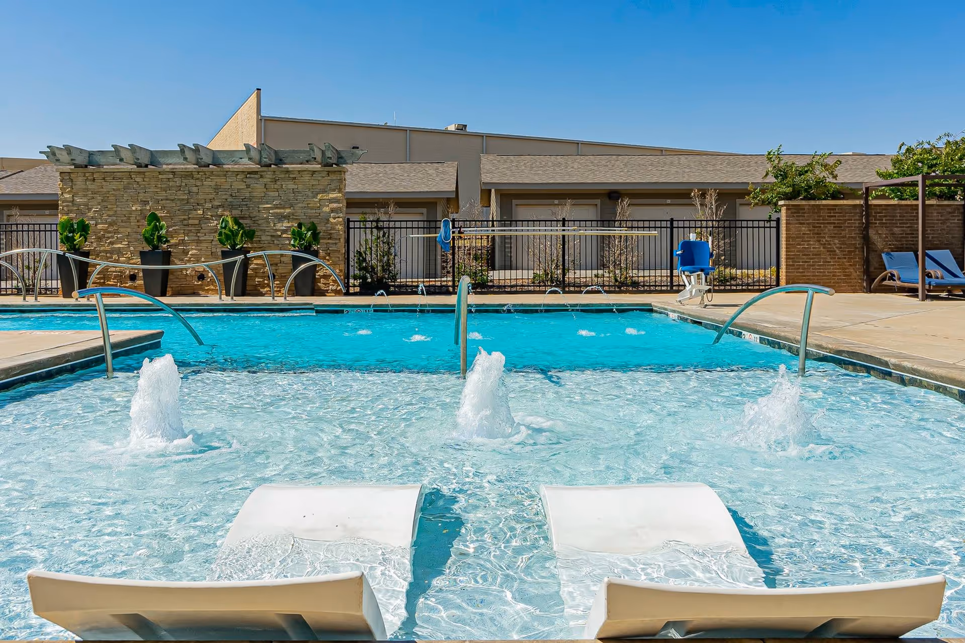 Outdoor swimming pool area with two white lounge chairs partially submerged in shallow water with small water fountains. The pool is surrounded by a concrete deck, black metal fence, and some potted plants. There are blue lounge chairs and a pool lift chair on the right side. The sky is clear and blue.