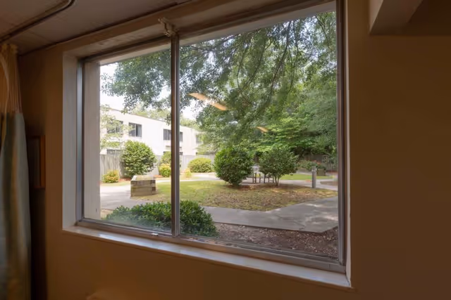 View through a large window showing an outdoor courtyard area with trees, bushes, paved walkways, and a building in the background.
