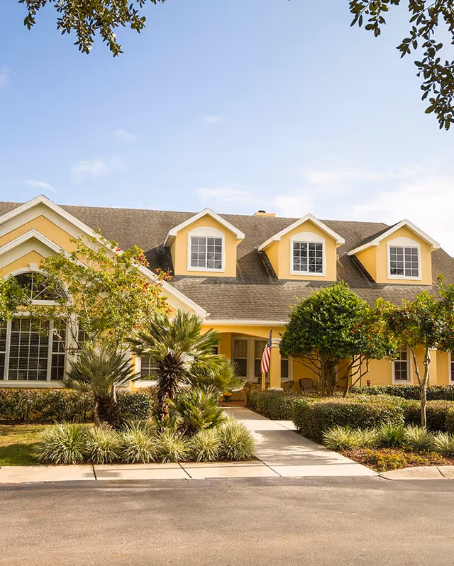 Front exterior view of a yellow two-story residential building with a gray shingled roof, three dormer windows, and a well-maintained garden with various shrubs and small trees. A concrete walkway leads to the entrance, where an American flag is displayed.