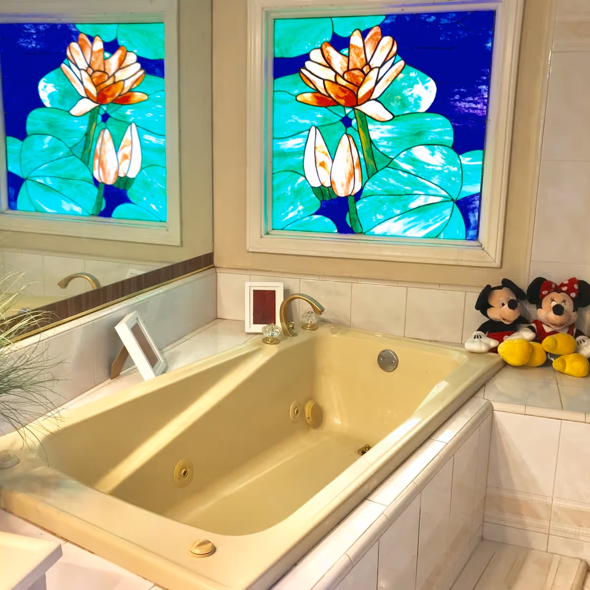 A beige bathtub with gold-colored faucet and crystal handles in a tiled bathroom. Above the tub are two stained glass windows featuring large green lily pads and white flowers. On the edge of the tub are two plush toys of Mickey Mouse and Minnie Mouse. A large mirror reflects part of the bathtub and a small framed picture is placed on the tub ledge.