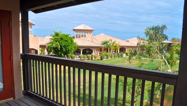 View from a balcony with wooden railing overlooking a green lawn and landscaped garden area with palm trees and shrubs. In the background, there are multiple buildings with terracotta tiled roofs under a blue sky with some clouds.