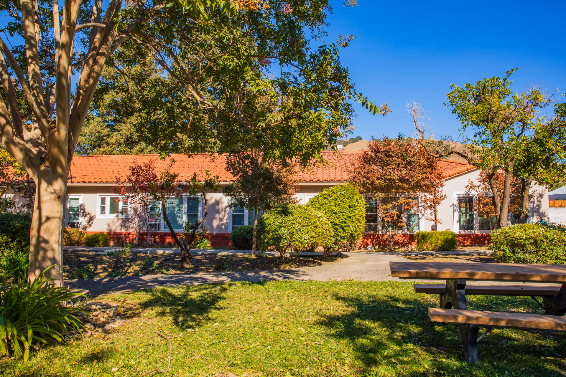 Front of a single-story building with a red tile roof, trees and shrubs in a sunny courtyard and a wooden picnic table in the foreground.