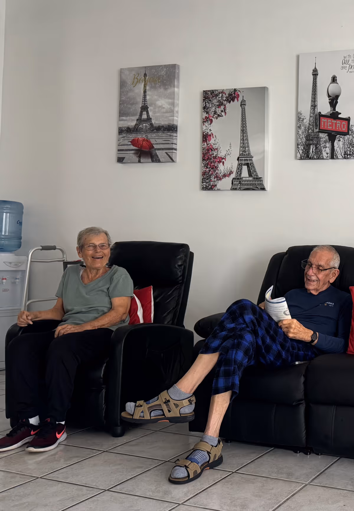 Two elderly people sitting and relaxing in black recliner chairs in a room with white walls. One person is smiling and wearing a green shirt and black pants, while the other is reading a booklet and wearing a dark long-sleeve shirt and blue plaid pants with sandals and socks. Behind them are three framed pictures of the Eiffel Tower on the wall, and a water dispenser is visible on the left side.