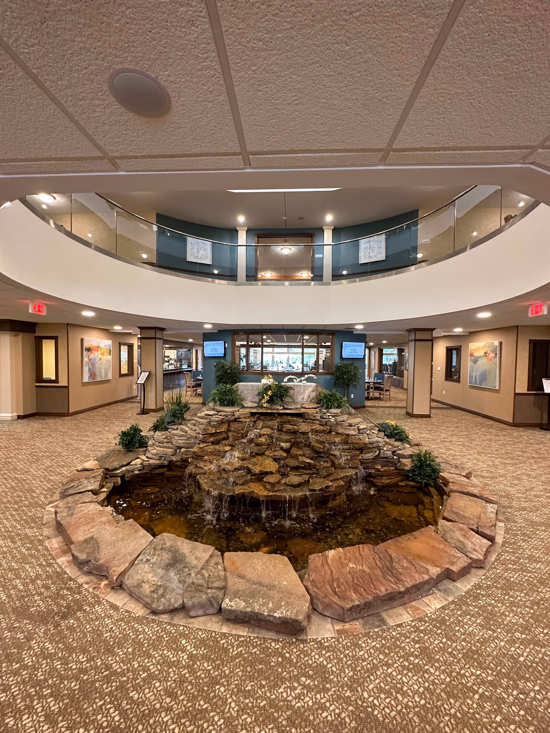 Interior view of a spacious senior living facility lobby featuring a central stone water fountain with cascading water surrounded by plants. The area has carpeted floors, beige walls with framed artwork, and a second-floor balcony with glass railing overlooking the lobby. There are exit signs and seating areas visible in the background.
