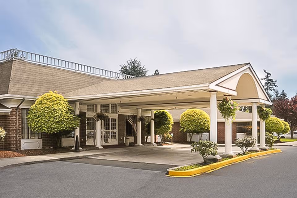 Exterior view of Hallmark Manor featuring a covered entrance with white columns, neatly trimmed round bushes, and a paved driveway under a partly cloudy sky.