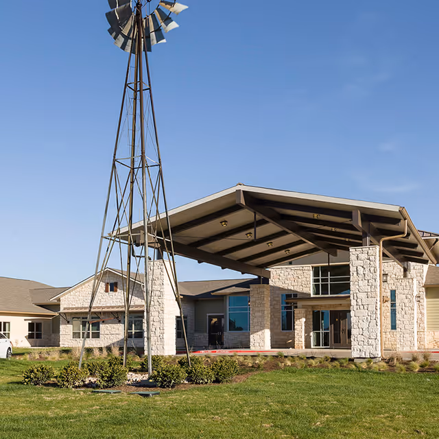 Exterior view of Lynridge of Waco Assisted Living & Memory Care building with a large covered entrance supported by stone pillars and a tall metal windmill structure on a grassy lawn under a clear blue sky.