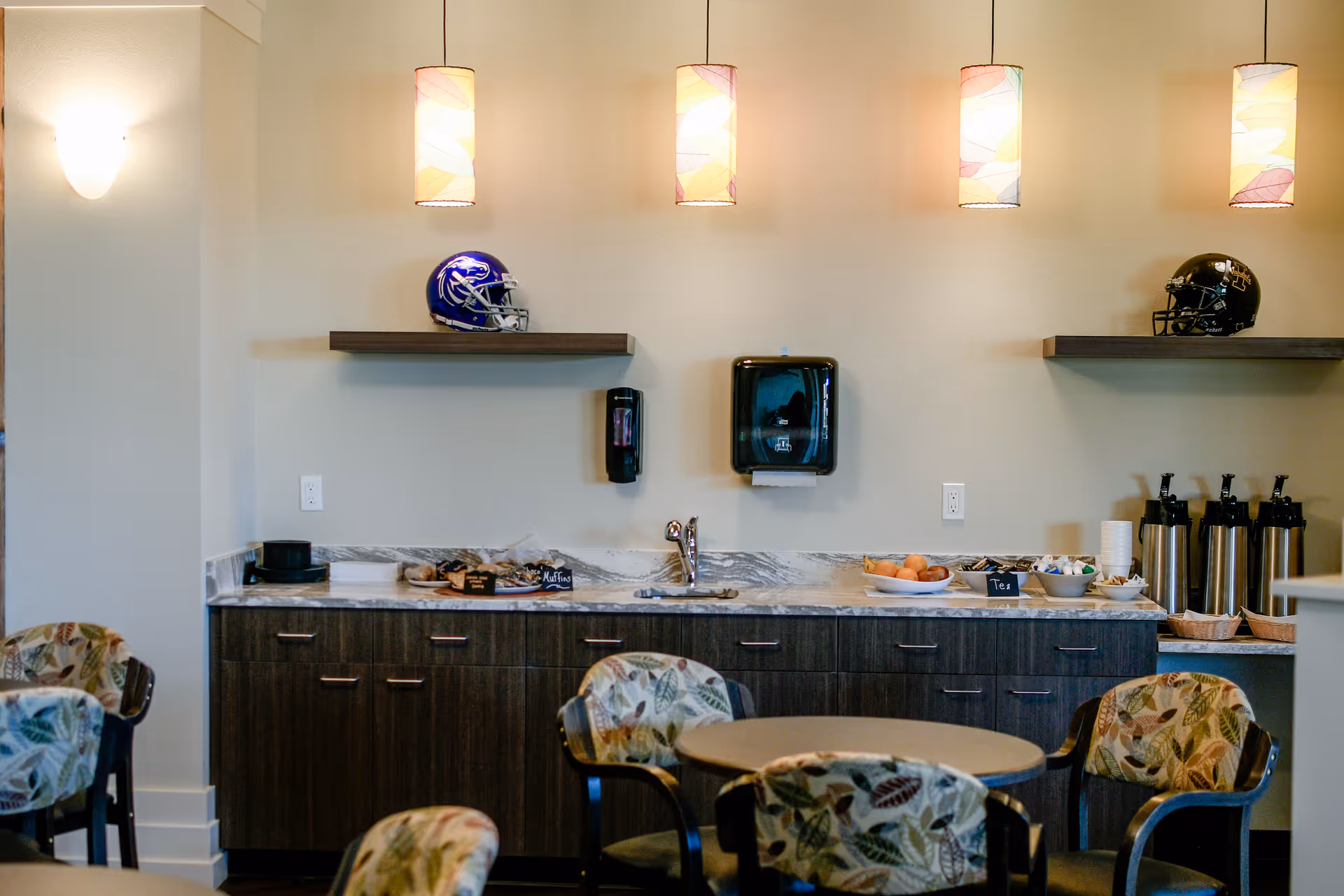 A cozy dining area with a round table and chairs featuring leaf-patterned upholstery. Against the wall is a countertop with a sink, coffee dispensers, a bowl of fruit, and trays with muffins and tea. Two floating shelves above the counter display football helmets. Three pendant lights hang from the ceiling, illuminating the space.