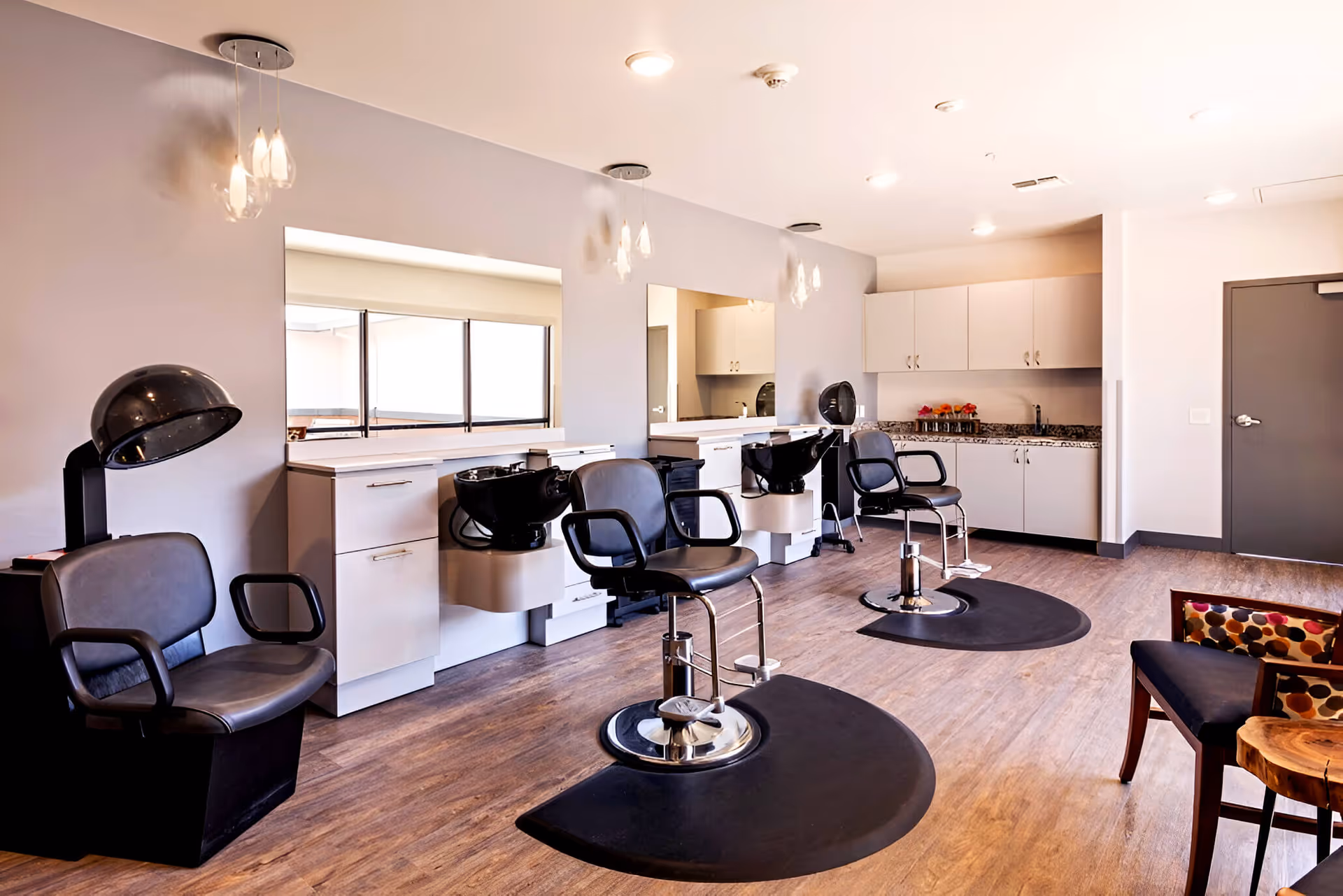 Interior view of a modern hair salon area with three black salon chairs, two black hair washing sinks, a hair dryer chair, large mirrors on the wall, white cabinetry, and wooden flooring. The room is well-lit with ceiling lights and pendant lights, and there is a seating area with chairs and a small table on the right side.