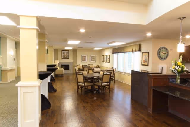 A spacious and well-lit common area with wooden flooring, multiple round tables with chairs, a fireplace at the far end, framed artwork on the walls, large windows with blinds, and a reception desk with a flower vase on the right side.