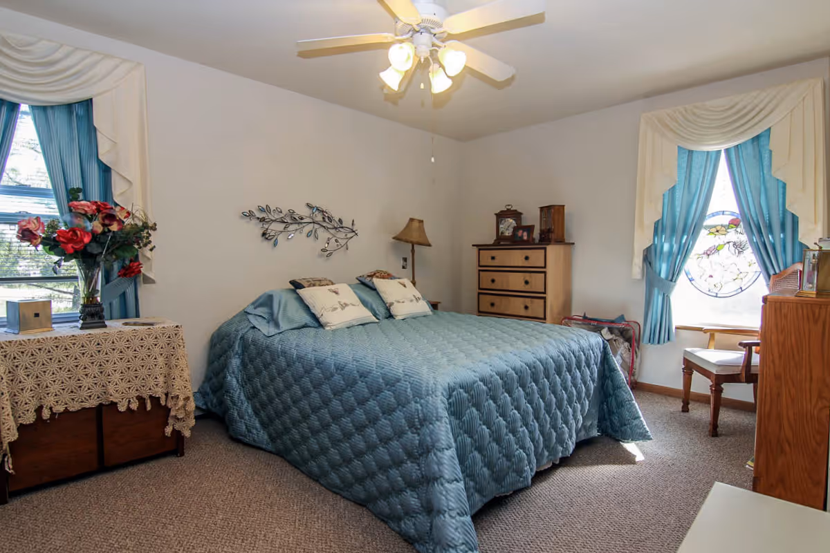 A cozy bedroom with a large bed covered in a blue quilted bedspread and two decorative pillows. The room features two windows with blue curtains and white valances, one with a stained glass design. There is a wooden dresser with a lamp and framed photos, a small table with a lace tablecloth and a vase of flowers, and a wooden chair near the window. A ceiling fan with lights is mounted on the ceiling.