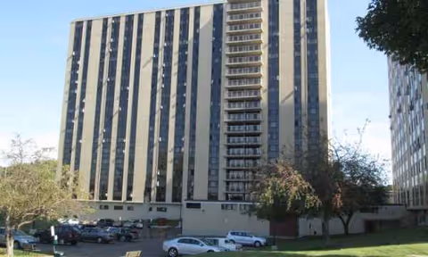 Tall gray high-rise apartment building with a parking lot in front and some trees.
