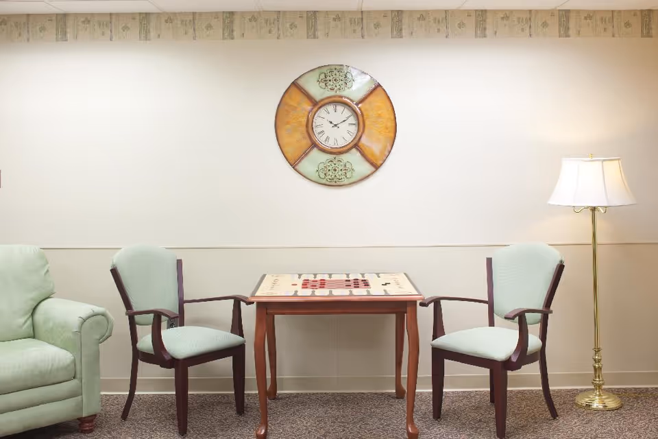 A small sitting area with two armchairs and a table set for a board game, a wall clock, floor lamp, and a sofa.