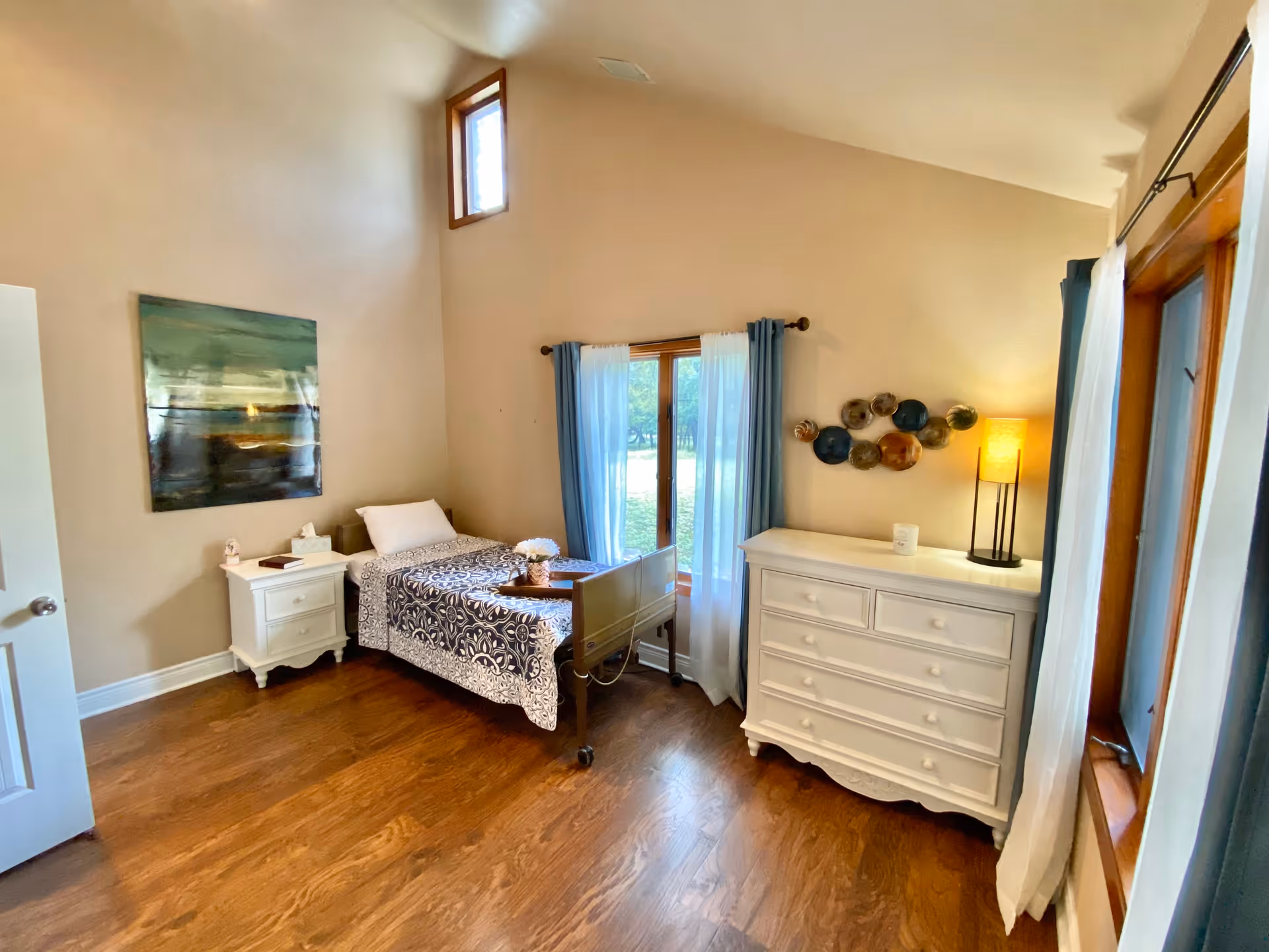 A bright and cozy bedroom with a single bed covered in a patterned bedspread, a white nightstand with a book and tissue box, a white dresser with a lamp and decorative items, and windows with blue and white curtains letting in natural light. The room has wooden floors and beige walls with a piece of artwork hanging above the nightstand.