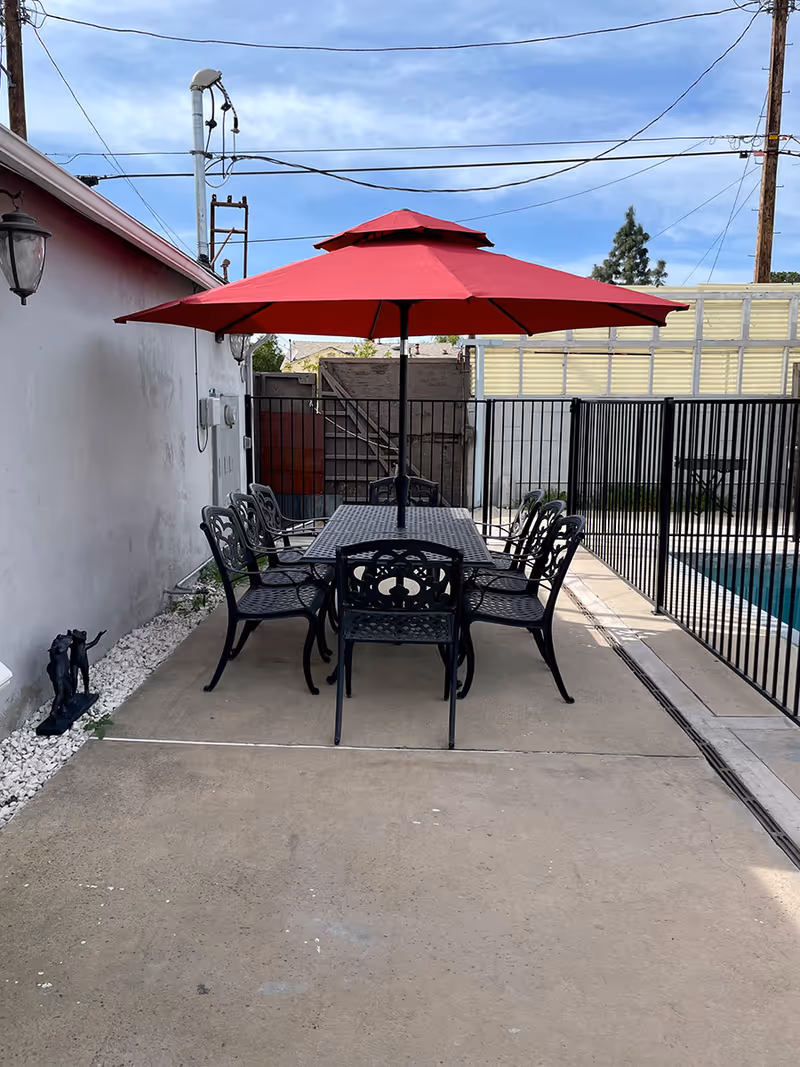 Outdoor patio area with a rectangular metal table and six matching chairs under a large red umbrella. The patio is next to a white wall with a lamp and some electrical boxes. A black metal fence surrounds a swimming pool on the right side. The sky is partly cloudy with visible power lines overhead.