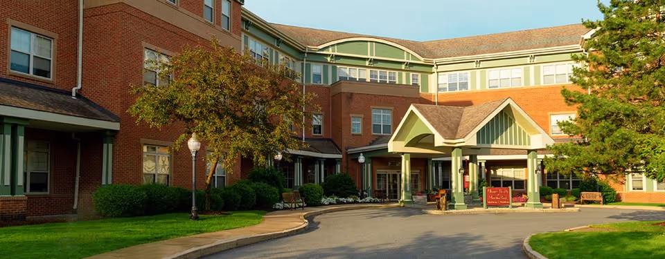Front entrance of a multi-story brick senior living facility with a covered porte-cochere, driveway, and landscaped grounds.