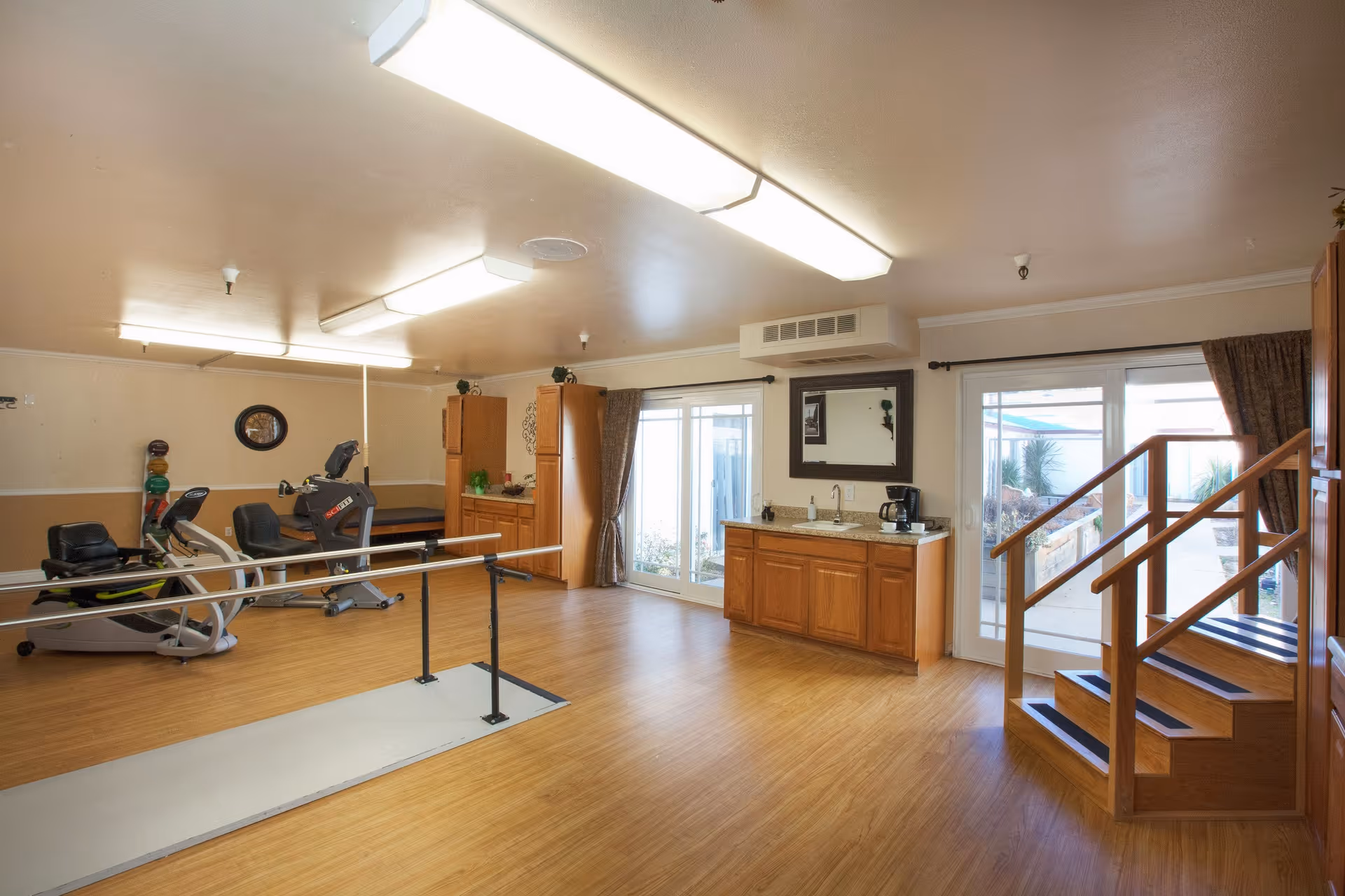 A spacious room with wooden flooring featuring exercise equipment including a recumbent bike and treadmill, parallel bars for walking practice, wooden cabinets, a countertop with a coffee maker, and a small staircase leading to a glass door that opens to an outdoor area.
