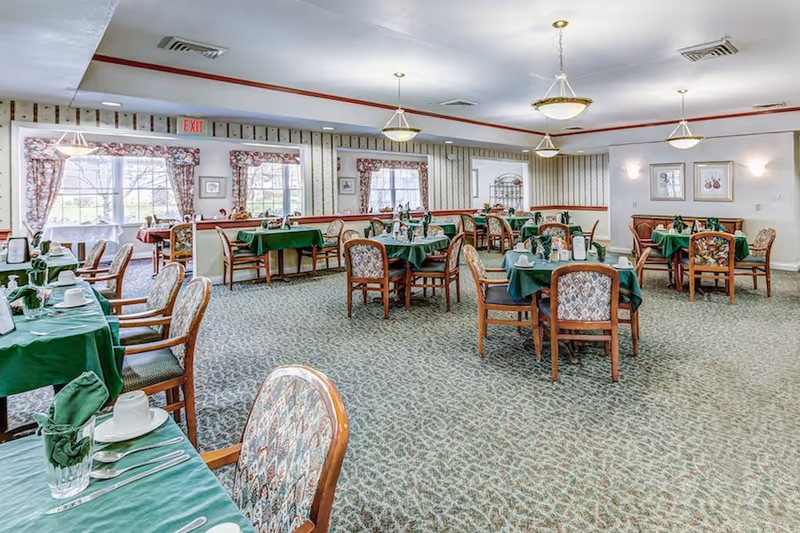 Spacious dining room with multiple tables covered in green tablecloths, wooden chairs, and place settings.