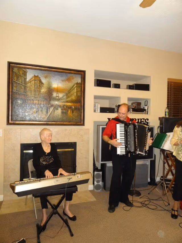 An elderly woman plays a keyboard while a man plays an accordion in a living room with a fireplace and framed painting.