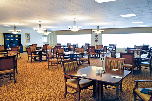 Spacious senior living dining room with multiple wooden tables and upholstered chairs, carpeted floor, and overhead chandeliers.