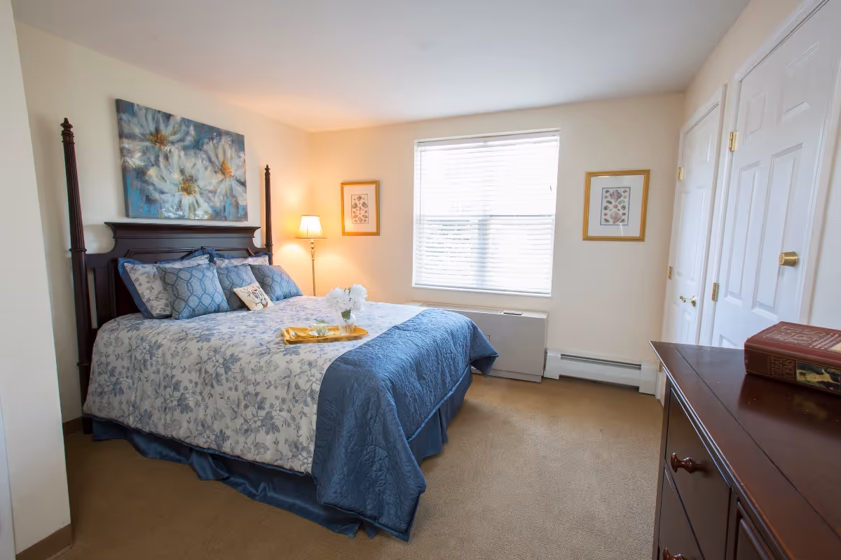 Bright bedroom with a large bed dressed in blue and white floral bedding, a bedside lamp, window with blinds, and a wooden dresser.