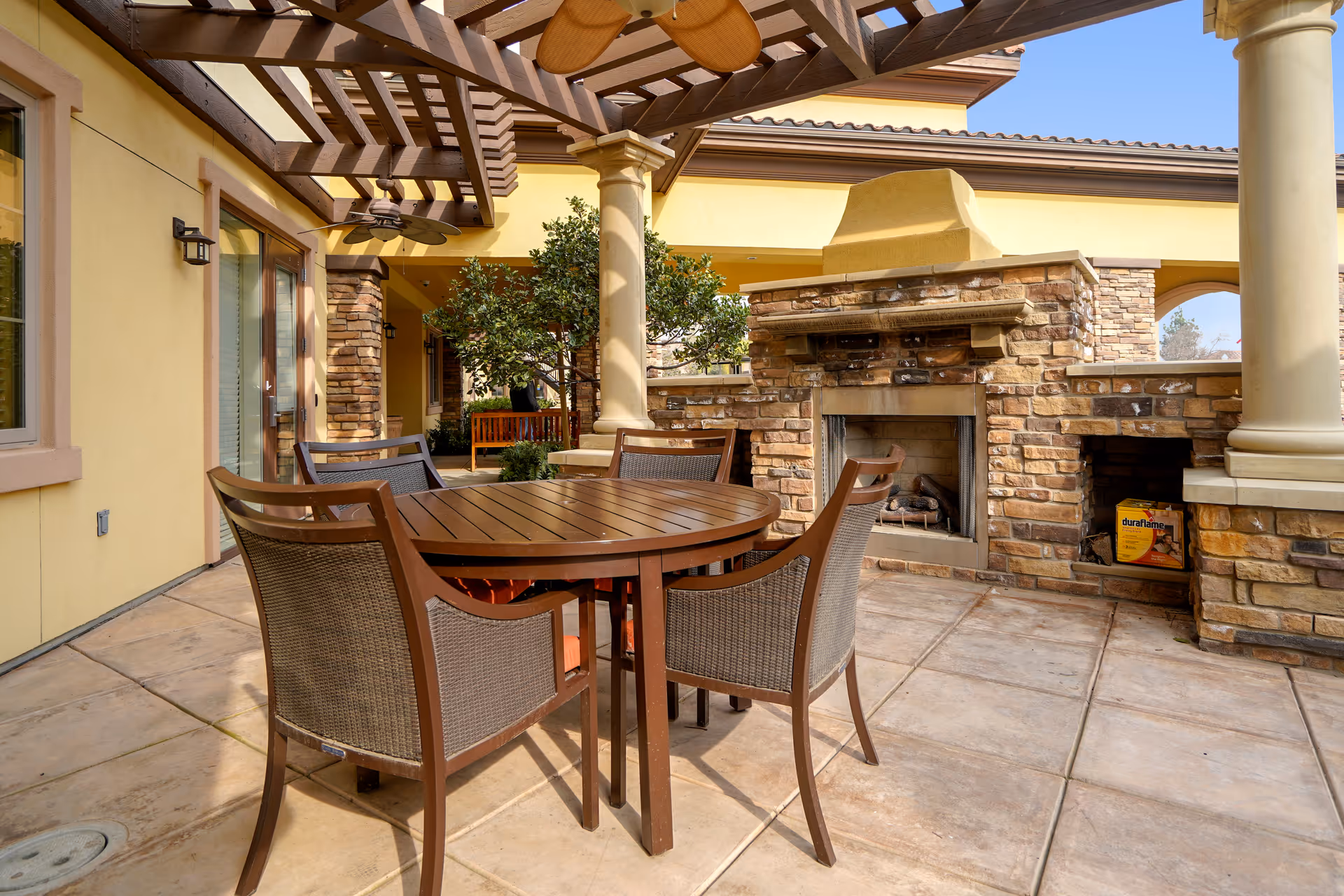 Covered outdoor patio with a round wooden dining table and chairs in front of a stone fireplace beneath a wooden pergola.