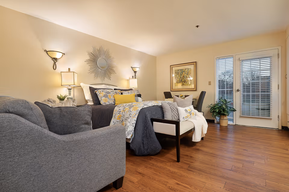 A cozy bedroom in an assisted living facility featuring a bed with floral and yellow accent pillows, a cushioned bench at the foot of the bed, a gray armchair, two bedside lamps, wall sconces, a decorative sunburst mirror above the bed, a framed painting on the wall, a small table with two chairs, a potted plant, and a door with window blinds leading outside. The room has wood flooring and soft yellow walls.