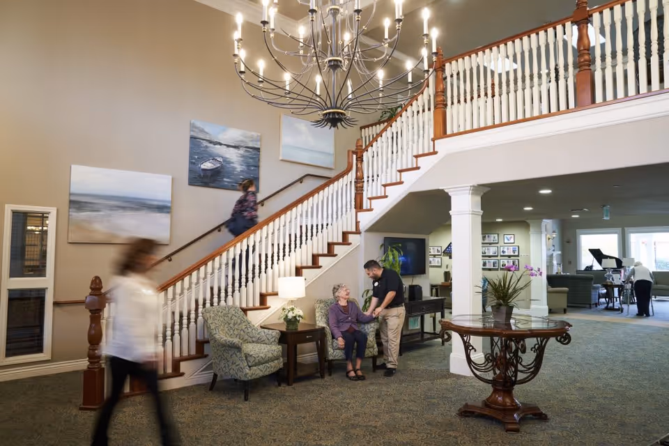 Lobby of a senior living facility with a grand staircase, chandelier, seating area, and staff assisting a resident.