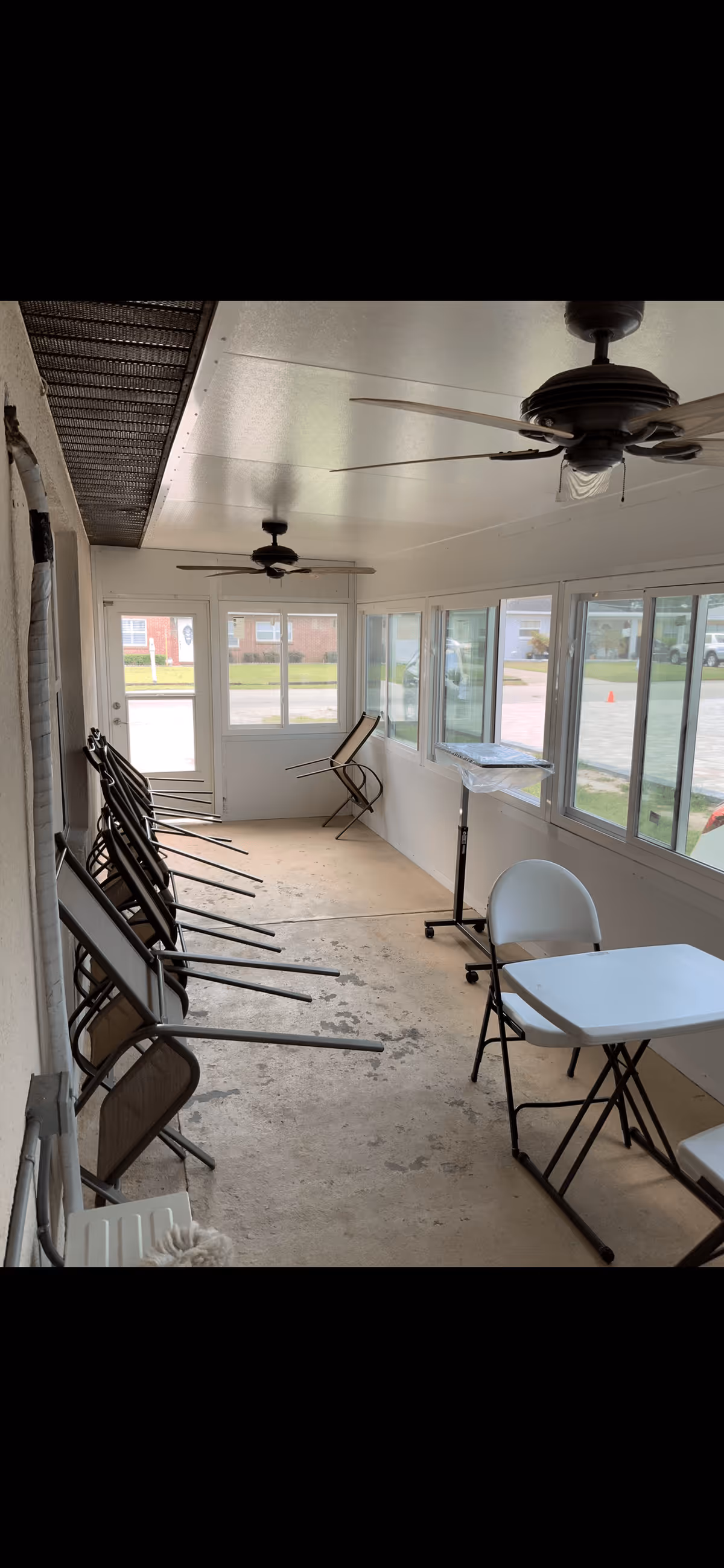 Enclosed sunroom/common area with stacked folding chairs, folding tables, and ceiling fans along a row of windows overlooking the street.