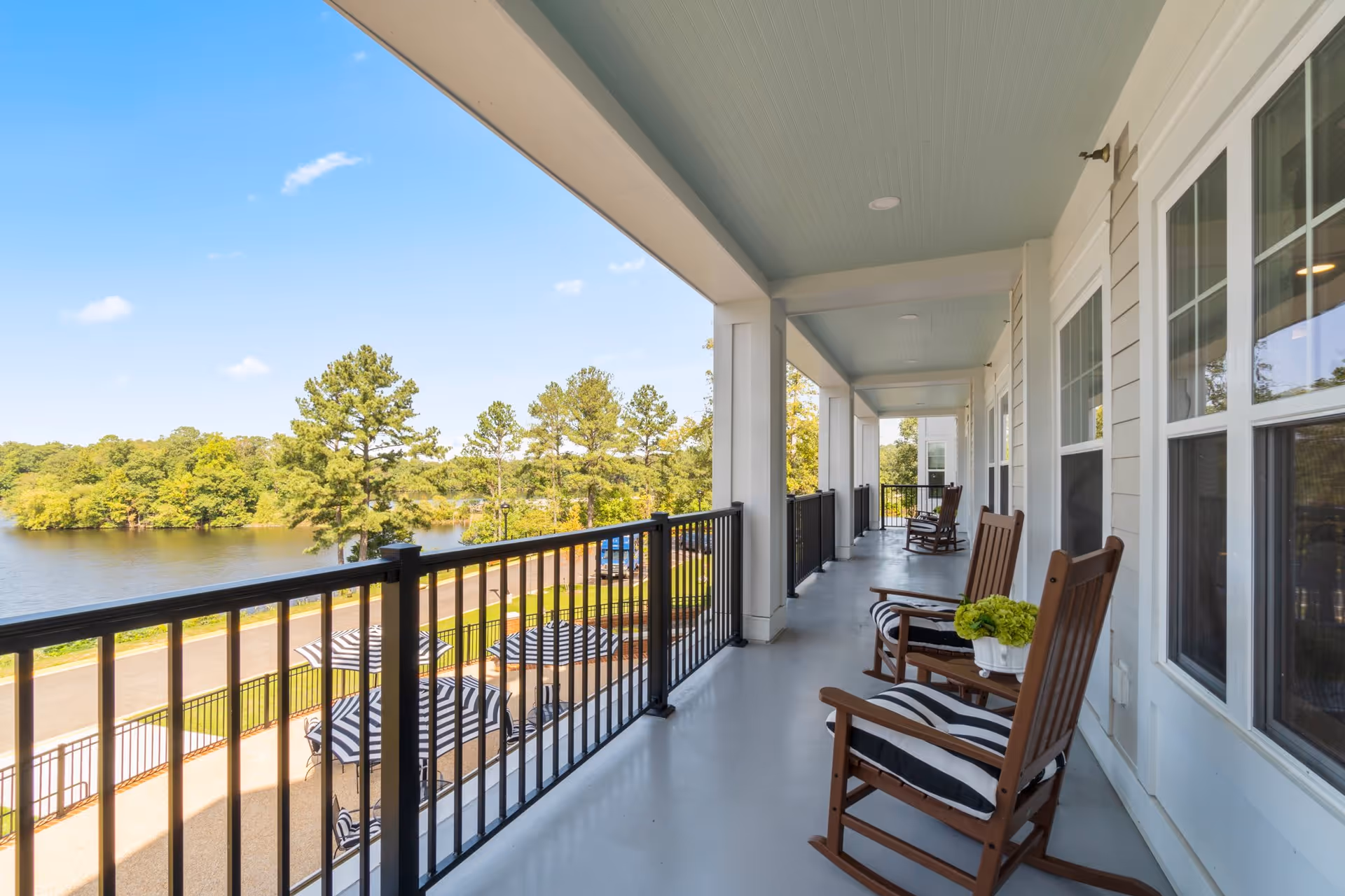 Covered balcony with wooden rocking chairs and small tables, overlooking a scenic view of a lake, trees, and outdoor seating with striped umbrellas.