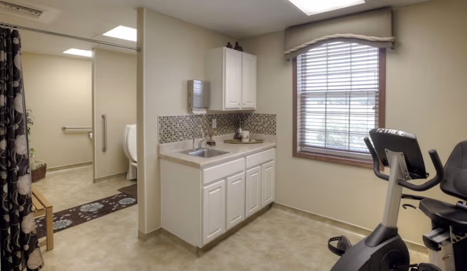 A small room with a white cabinet and sink, mosaic tile backsplash, a window with blinds, and an exercise bike. To the left, there is an open doorway leading to a bathroom with a toilet, grab bars, and a shower curtain.