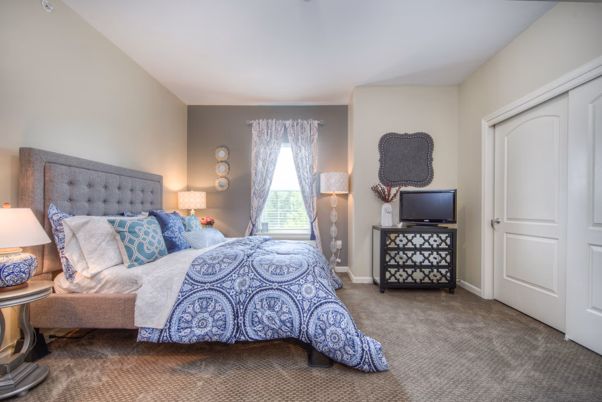 A neatly made bed with a tufted gray headboard and blue patterned bedding in a senior living bedroom. The room features a window with patterned curtains, two bedside lamps, a decorative chest of drawers with a small TV on top, and a carpeted floor.